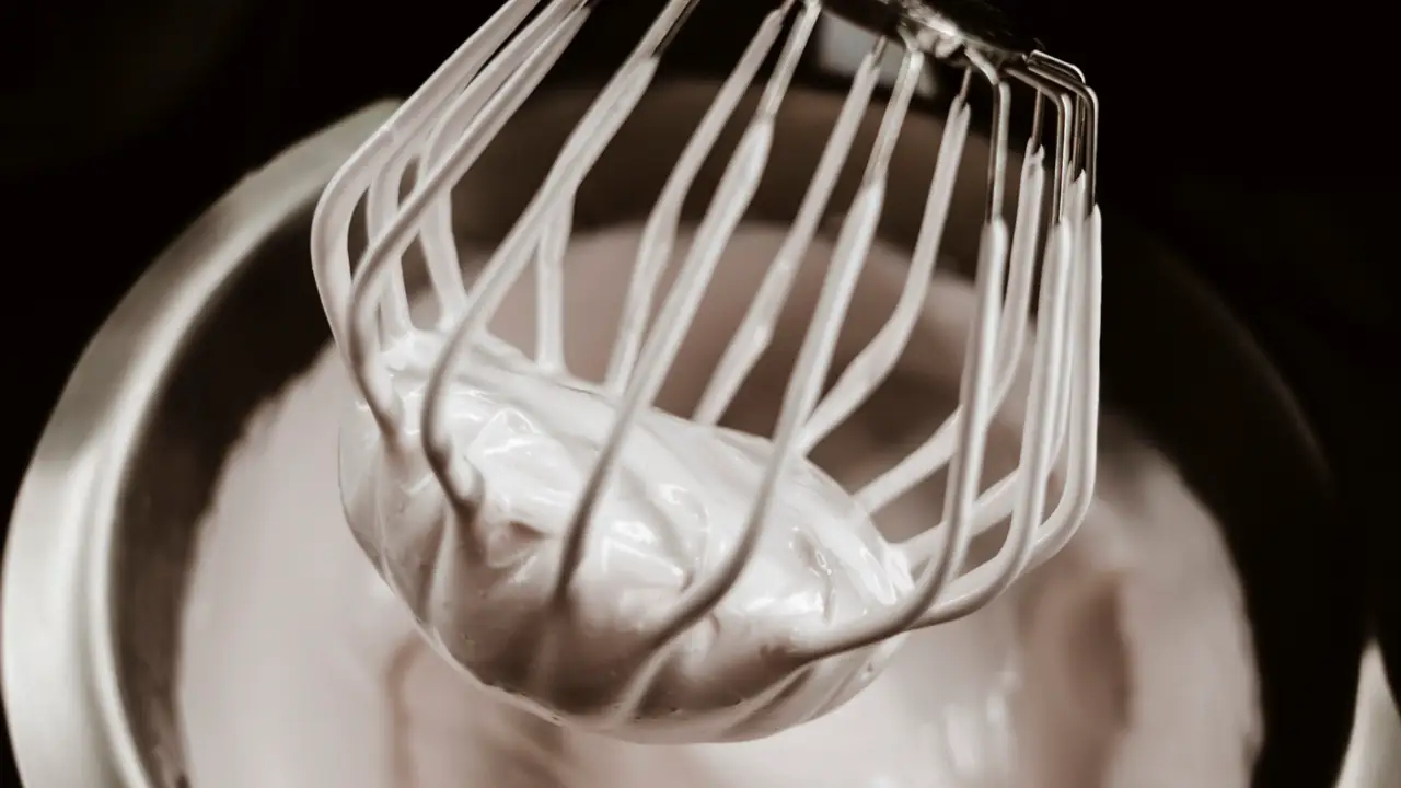 A mixer whisk in a metal bowl of white whipped egg whites